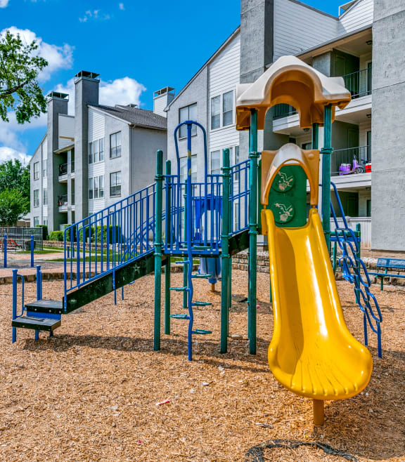 a playground with a yellow slide and blue playset in front of apartments