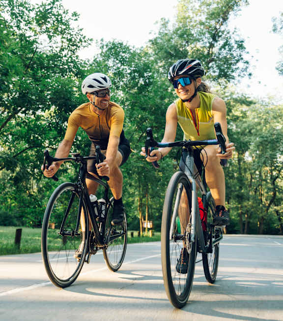 Two cyclists wearing helmets and sunglasses ride their bikes on a road.