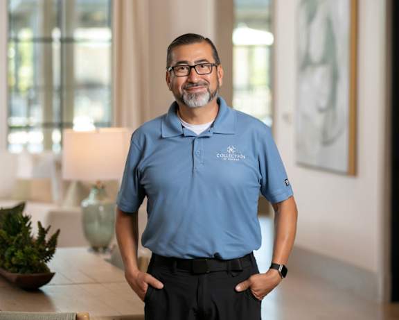 A man in a blue shirt and beige pants stands in a room with a table and a plant.