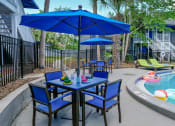 Thumbnail 14 of 23 - Blue patio tables and chairs with blue umbrellas next to community pool in Mandarin Bay apartments in Jacksonville, Florida.