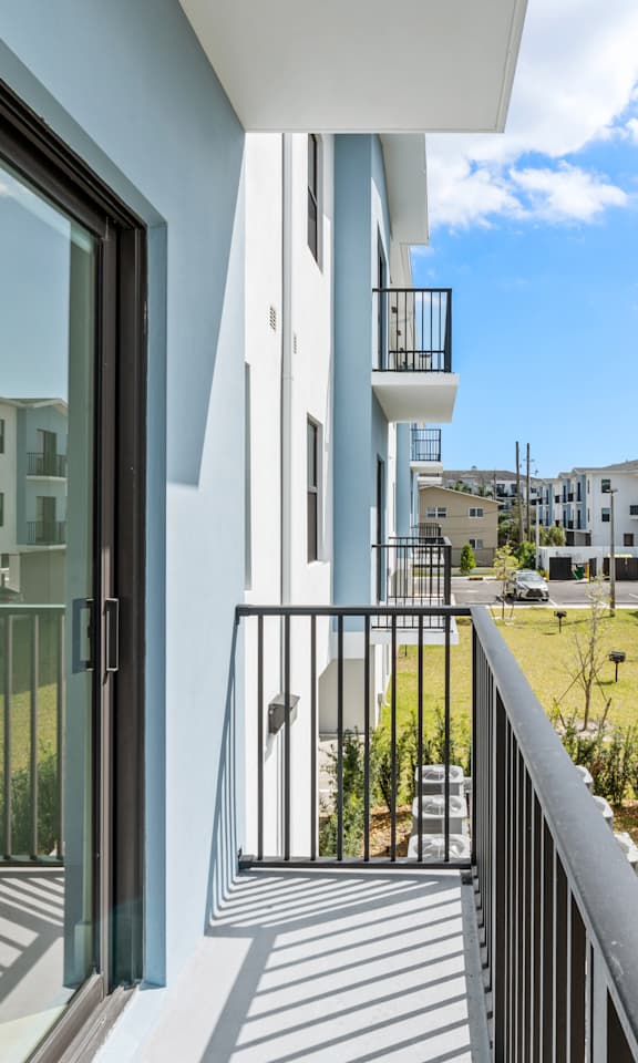 A balcony with a glass door overlooks a grassy area with other buildings in the distance.