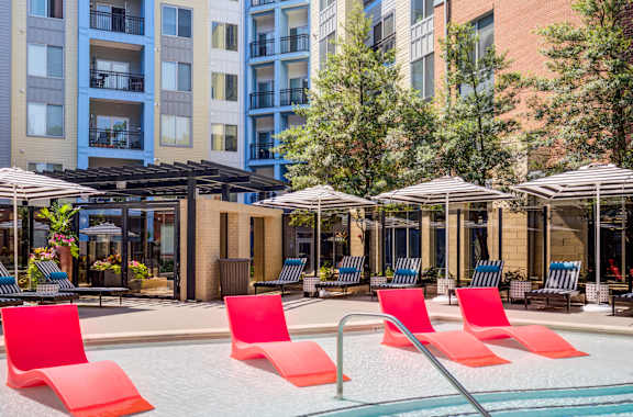 A pool with red chairs and umbrellas in front of apartment buildings.