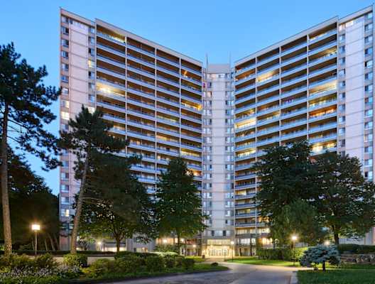 A modern building with a symmetrical design is illuminated from within and surrounded by trees.