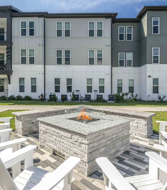 A modern outdoor seating area with white chairs and a fire pit in front of a multi-story apartment building.