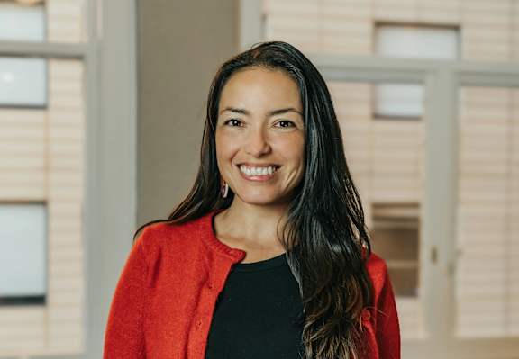 A woman with long dark hair wearing a red cardigan and black top standing in front of a window.