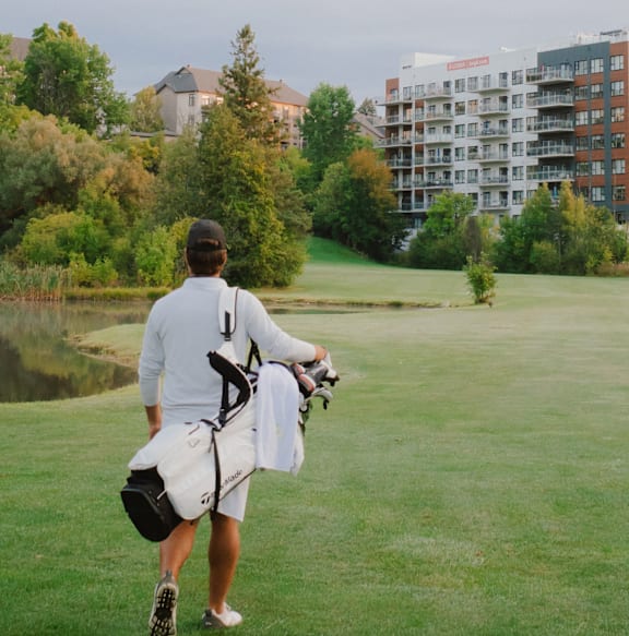 A man in a white shirt and shorts is walking on a golf course carrying a golf bag.