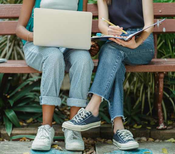 Two people sitting on a bench with one person using a laptop and the other writing in a notebook.