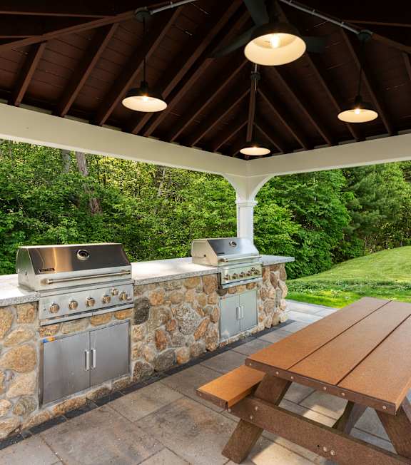 A wooden picnic table is in front of a stone wall with a grill and a sink.