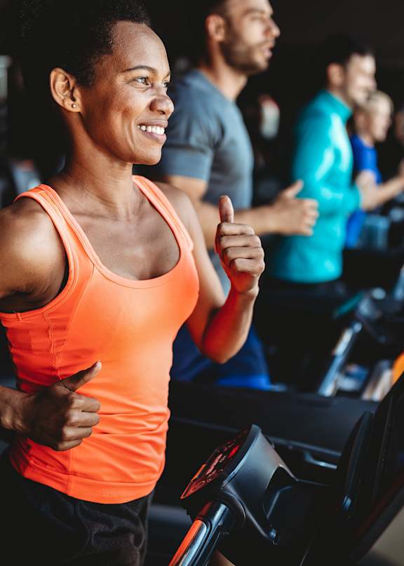 A man in a white t-shirt is running on a treadmill in a gym with other people exercising around him.
